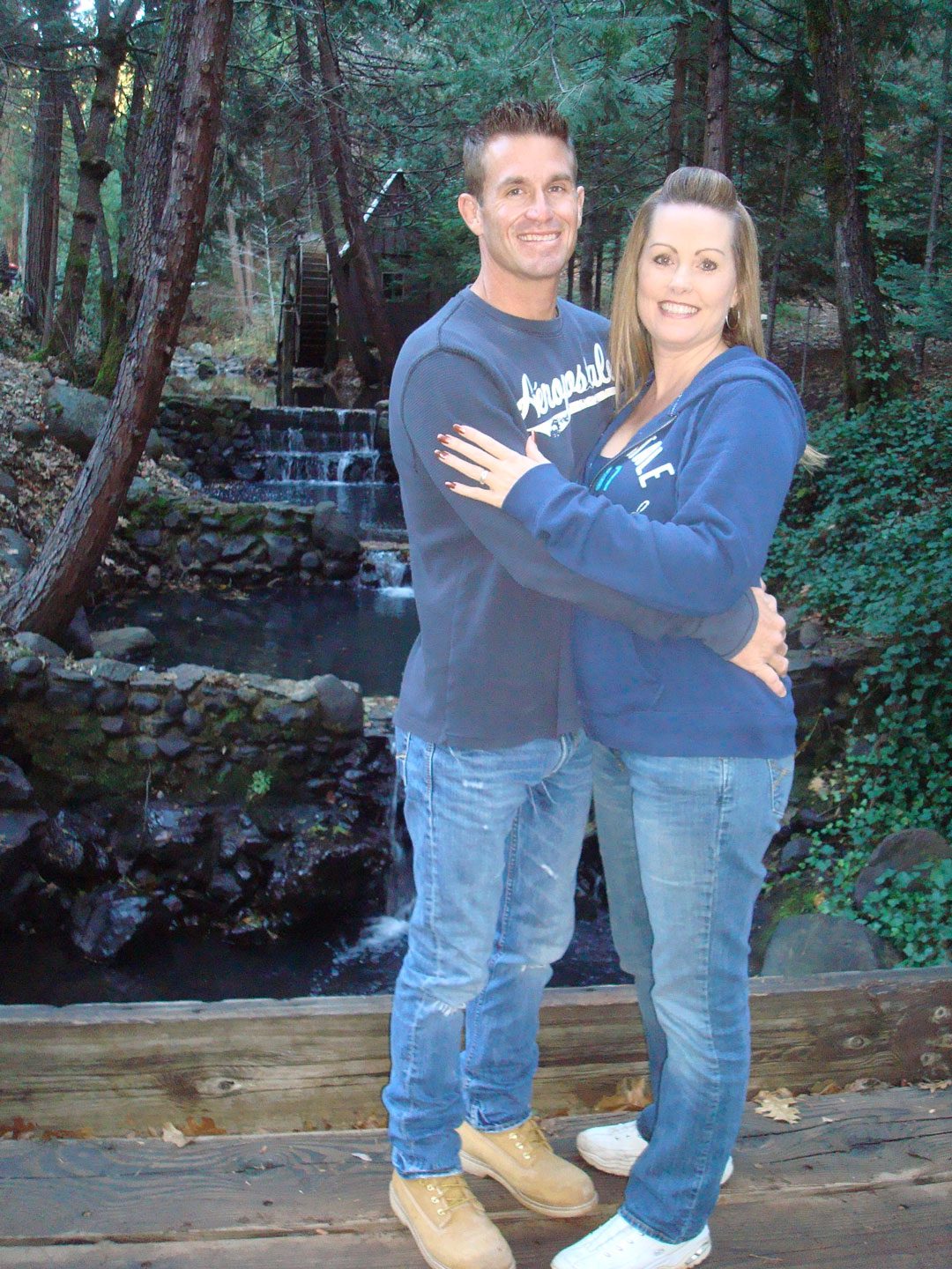 dave_and_marcia A man and a woman standing close together and smiling in front of a small waterfall and wooden structure, surrounded by trees and greenery.